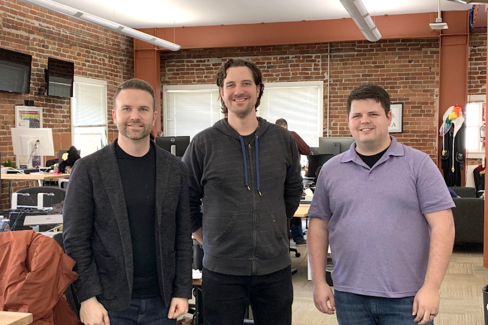three men standing in an open office space, smiling at the camera
