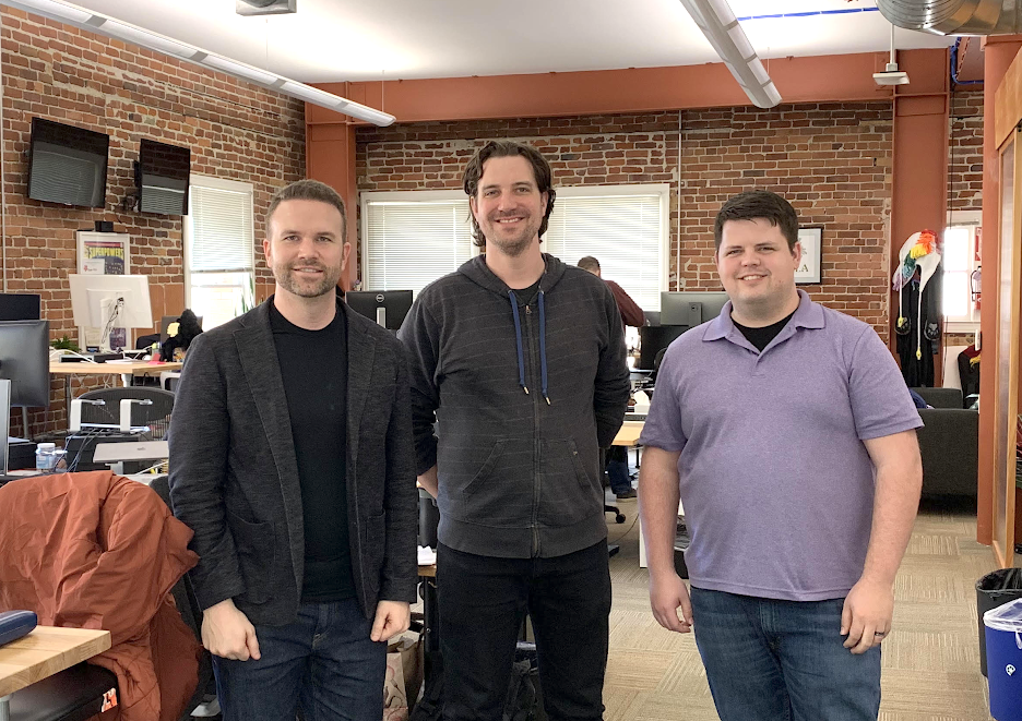 three men standing in an open office space, smiling at the camera